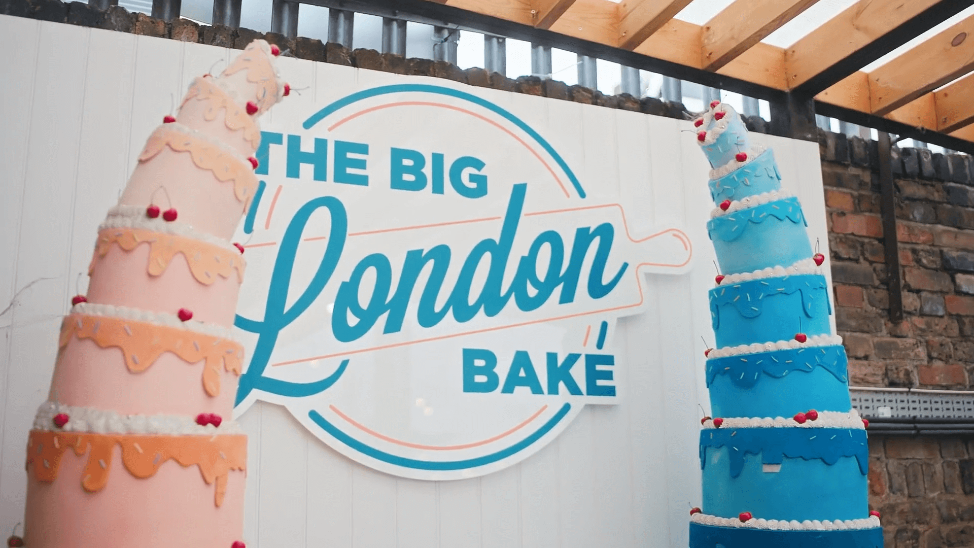 The Big London Bake logo between two tall decorative cakes in pink and blue with dripping icing and cherry decorations
